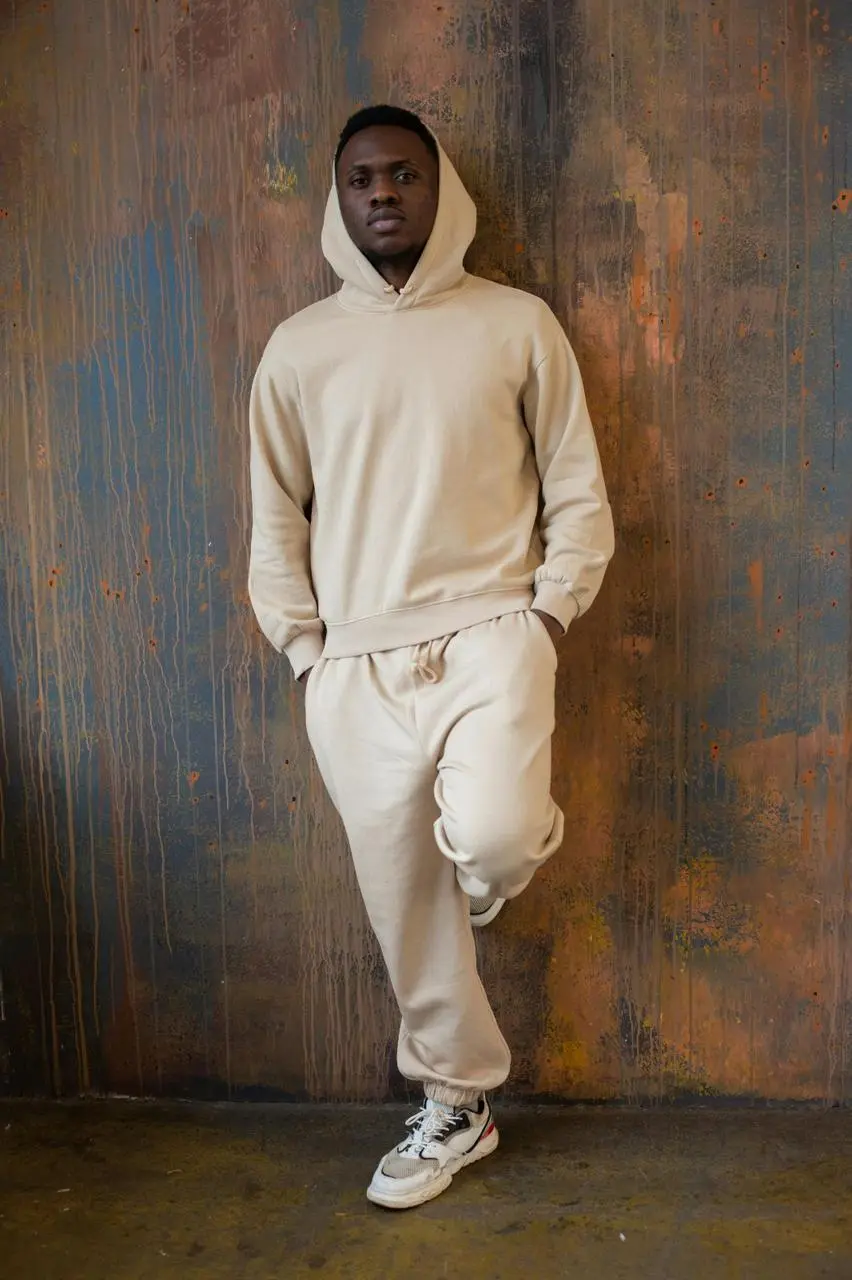 A man dressed in a beige sweatshirt and sneakers poses, reflecting a comfortable and casual outfit choice.