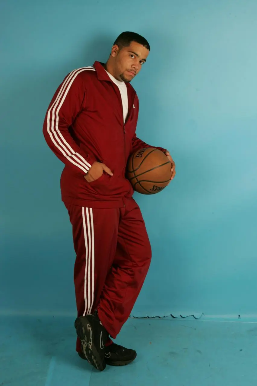A man in a red track suit stands holding a basketball, ready to play on an outdoor court
