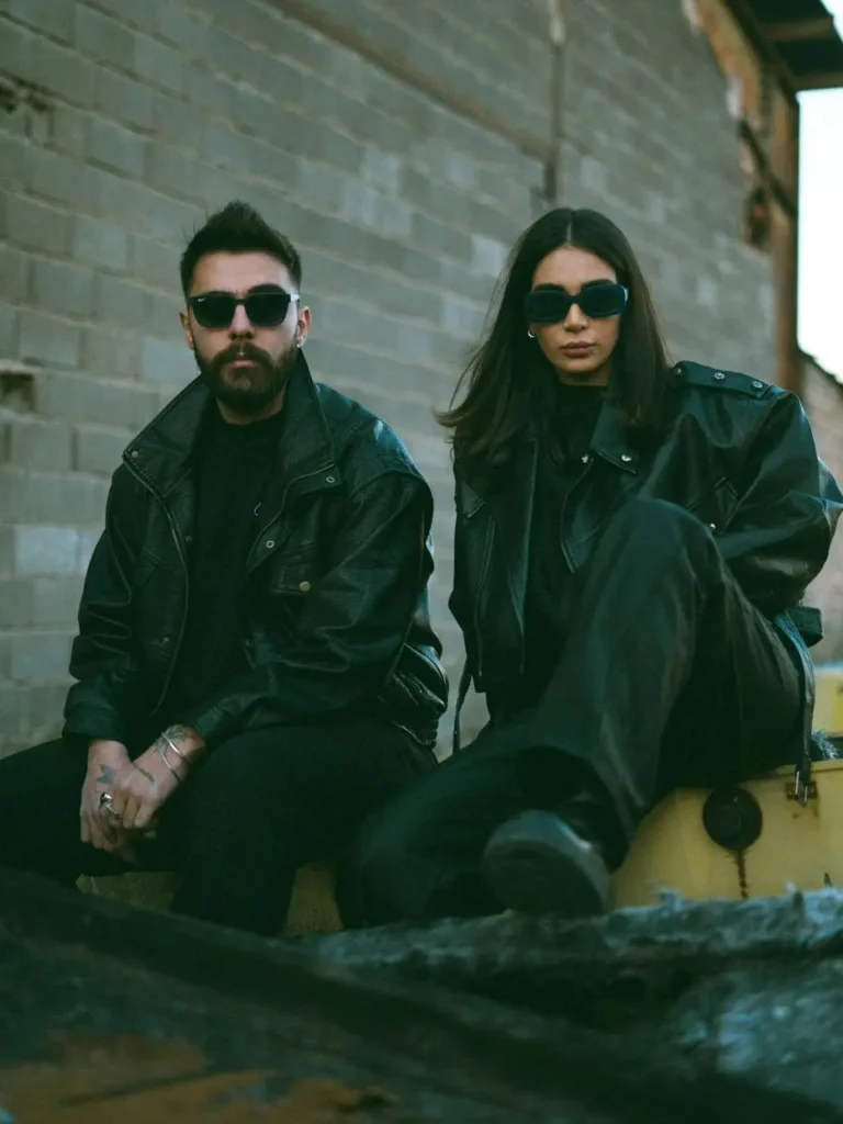 A pair of people wearing black leather jackets sitting together on a bench, appearing relaxed.